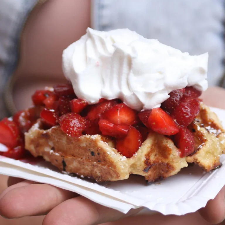 The image depicts a person holding a plate with a serving of waffle topped with fresh, sliced strawberries and a generous dollop of whipped cream. The whipped cream's fluffy texture suggests it might have been created using a nang, a culinary tool used for making whipped cream and other aerated food items. This scrumptious scene highlights the delightful result of one of the nangs' features, which is to effortlessly top desserts with perfectly whipped cream.