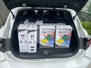 Open trunk of a car filled with numerous boxes of 'ignite' nitrous oxide, each box marked with handling instructions "THIS SIDE UP" and hazardous material warnings, indicating careful transport of sensitive materials.
