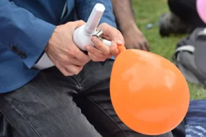 A person inflating an orange balloon using a manual air pump. The individual is sitting down, wearing a blue jacket and grey pants. There's grass in the background, suggesting this activity might be taking place outdoors, possibly at a park or an outdoor event.