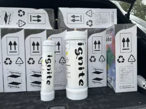 The image shows several cylindrical canisters and boxes labeled "ignite" in a car's trunk. These canisters are typically known as cream chargers. They are usually used with a whipped cream dispenser to aerate the cream with nitrous oxide, which is what the label "N2O" on the canisters stands for. Cream chargers are widely used in culinary applications to create whipped cream for desserts, beverages, and other dishes. The boxes also have recycling symbols and directions indicating that they are made of steel ("FE") and offer information on proper handling and disposal.